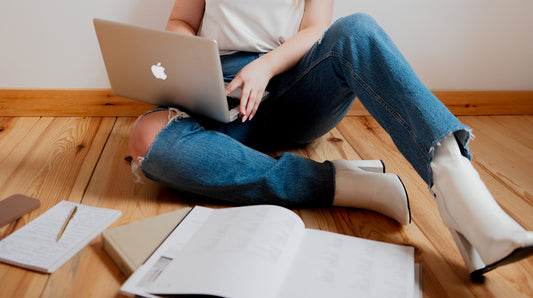 person sitting on wooden floor with mac laptop surrounded by books and notepads