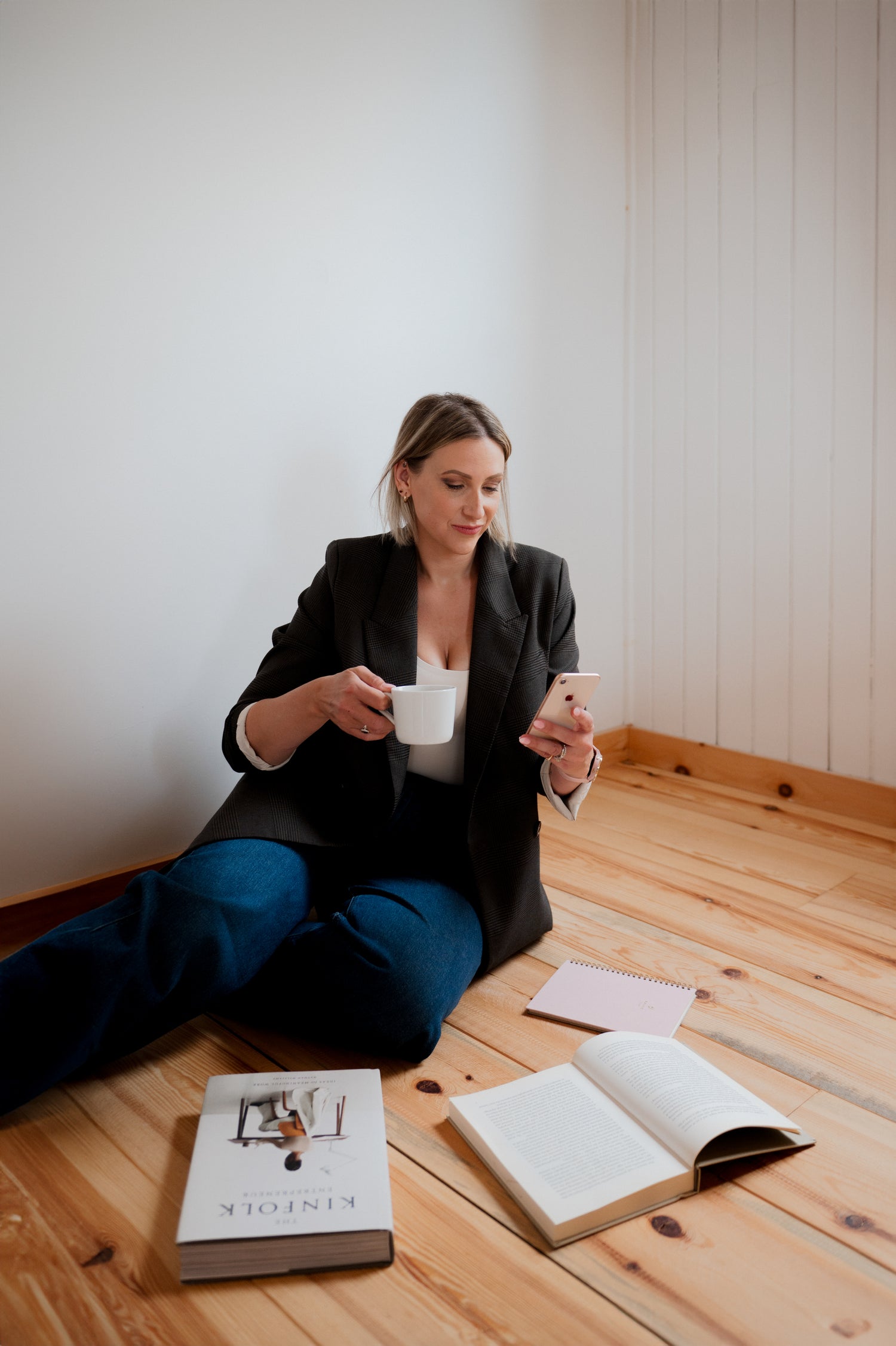 Blonde woman, Sheena Switzer, Blonder Marketing sitting on wooden floor holding coffee and cell phone with books in front of her